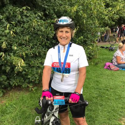 image of supporter Sue standing next to her bike wearing a white T shirt and helmet. Has a blue ribboned metal around her neck.