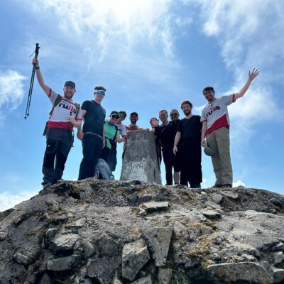 Image of 10 people at the summit of Ben Nevis in Scotland as a part of the three peaks challenge. They are standing at the stone at the top and two people have their hands in the air. Bright blue sky background.