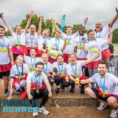 Group photo of the participants in the rough runner challenge event. All are wearing white t shirts and red tutus with their hands in the air. They have medals on a blue ribbon around their necks.