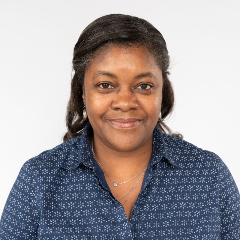 Image of a black woman with dark hair and brown eyes wearing a navy blue button up top with a flower pattern and two silver necklaces.
