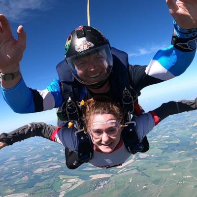 Image of Rachelle, a white woman with brown hair, tandem skydiving with a white man wearing a helmet. They are mid-air with their arms out in front of them.