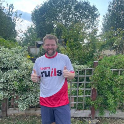 Image of rules supporter Ashley standing in front of a hedge in a garden wearing a team Ruils T shirt as he prepares for his skydive.