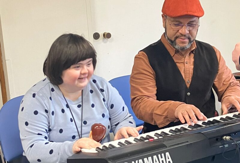 Image of a man wearing an orange flap cap and glasses seated at a piano next to a young woman with Down syndrome wearing a grey and black polka dot top. They are both playing the piano with their hands.