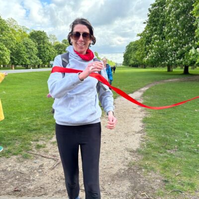 Image of a woman crossing the red ribbon finish line of the London Marathon my way marathon walk. She is wearing brown sunglasses and a grey hoodie.