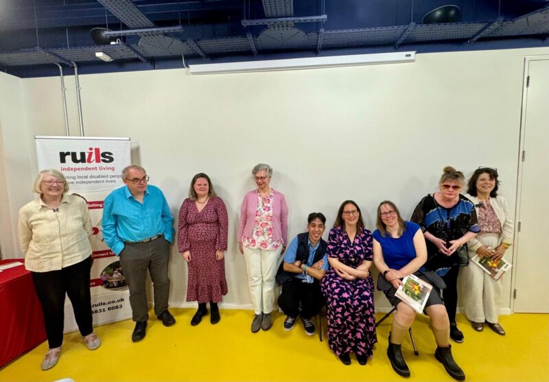 Group photo of the rules creative writing group at the launch of their creative writing anthology they are standing at the front of the room in front of a banner and the wall behind them is white with a yellow floor.