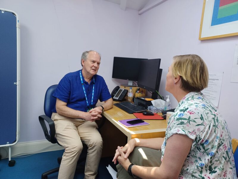 Image of Social Prescribing Link Worker James, sitting at a desk in a GP surgery, speaking to a woman sitting across from him.