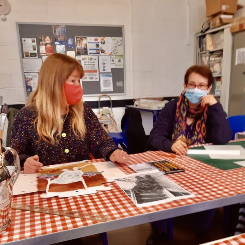 Two members of the art group seated at a table with a red gingham tablecloth. They are both wearing masks and in front of them on the table are various art supplies.