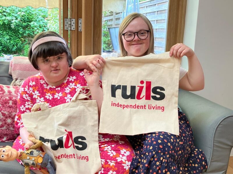 Two young women with Downs Syndrome sitting on a green couch holding their Ruils tote bags.