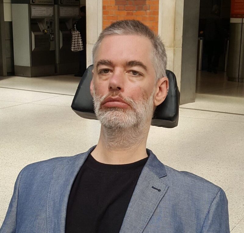 A headshot of previous trustee Alan Benson Wearing a dark grey suit and black T shirt sitting in his wheelchair.