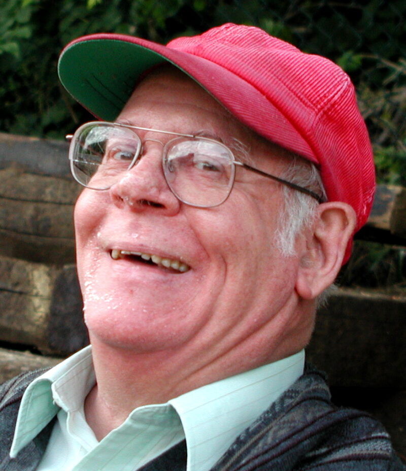Headshot of founding member Alan Pinn wearing a red baseball cap and light green button shirt.