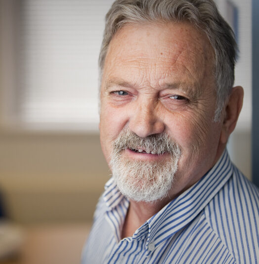 Image of Bob Burgess founding member of Ruils wearing a blue and white striped button shirt. He is a white man with grey hair and a grey and white goatee.