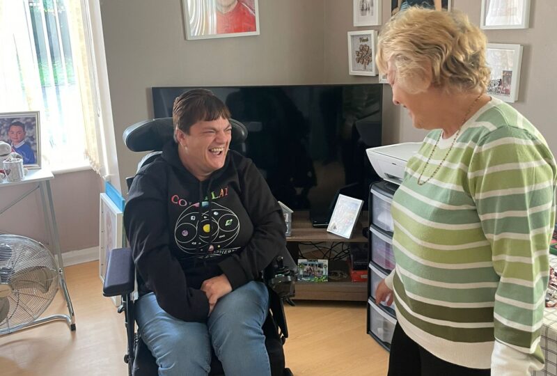 Image of a direct payment client Shirley who is seated in her wheelchair With her personal care assistance standing next to her. They are laughing about something that has been said. They are in the client's home.