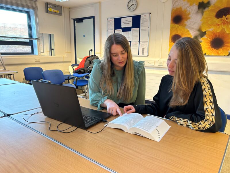 Image of pathways benefits advice case worker Emily sitting next to her client at a table in a meeting room. They are looking at a book that is sitting on the table next to a black laptop.