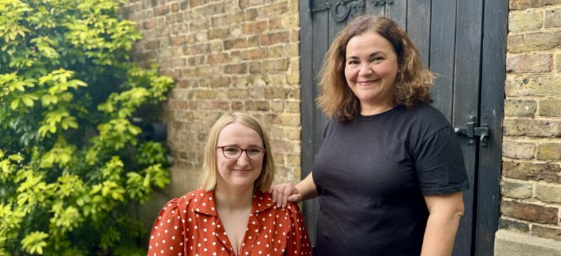 Image of Carla and Josie who are Ruils independent living advisers, standing in front of a brick wall and a wooden door both are looking and smiling at the camera.