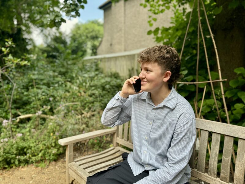 Image of a woman with short brown hair sitting on a wooden bench holding up a mobile phone to her ear