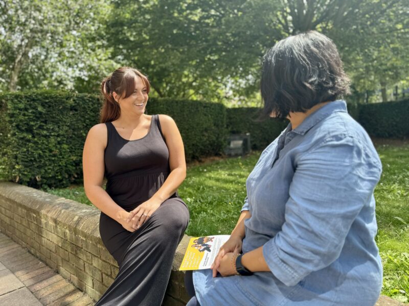 Two women sitting on a brick wall in a town centre speaking with each other. One is holding a Ruils leaflet and the other is listening and smiling.