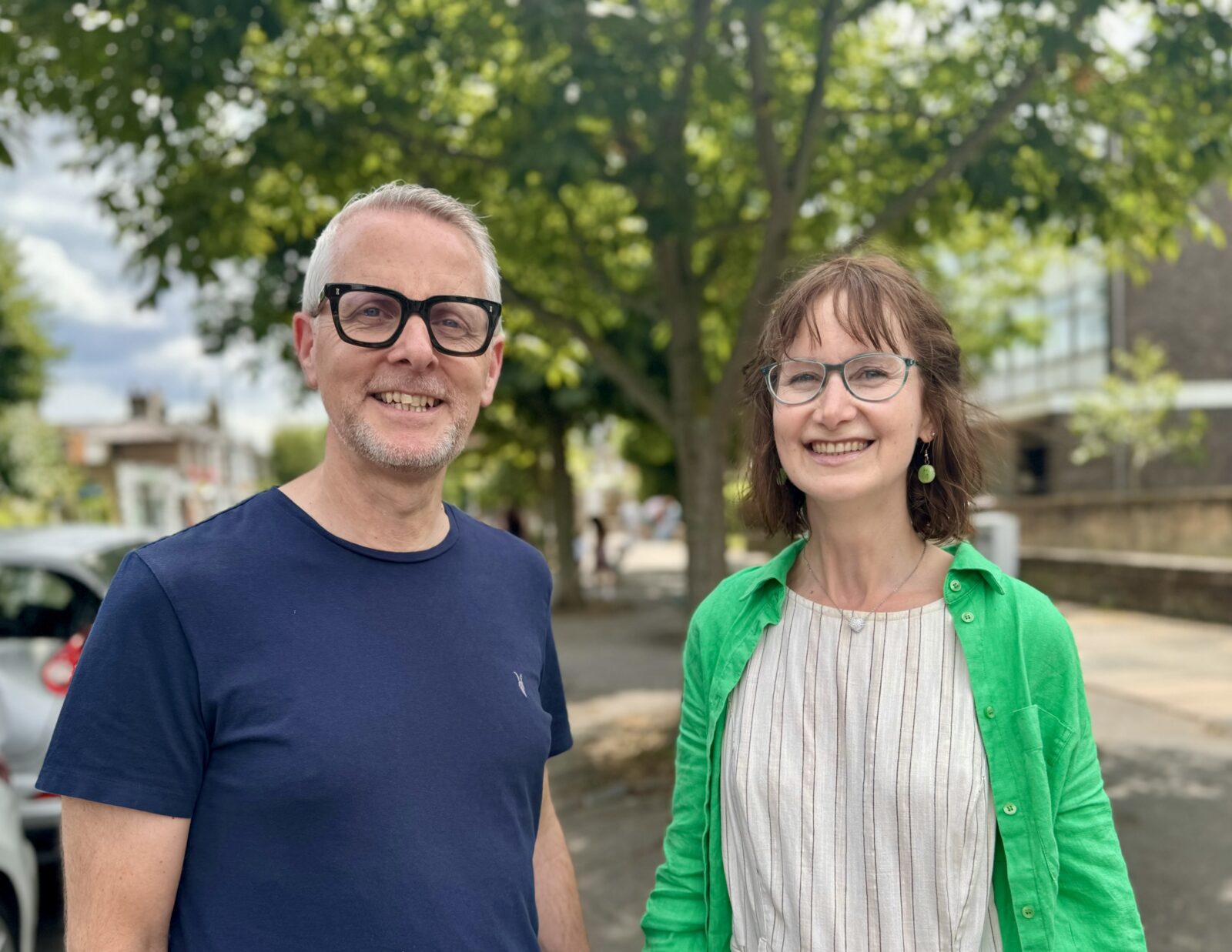 Community health and well being workers John and Susie standing on the pavement in front of a tree with green leaves. Susie is a white woman wearing a tan dress and green top and Phil is a white man wearing a navy blue T shirt and dark glasses.