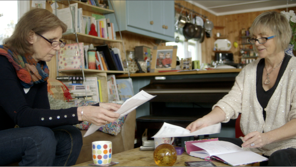 Two women sitting next to a coffee take looking at paperwork. Both are wearing glasses and have a cup of tea in front of them.