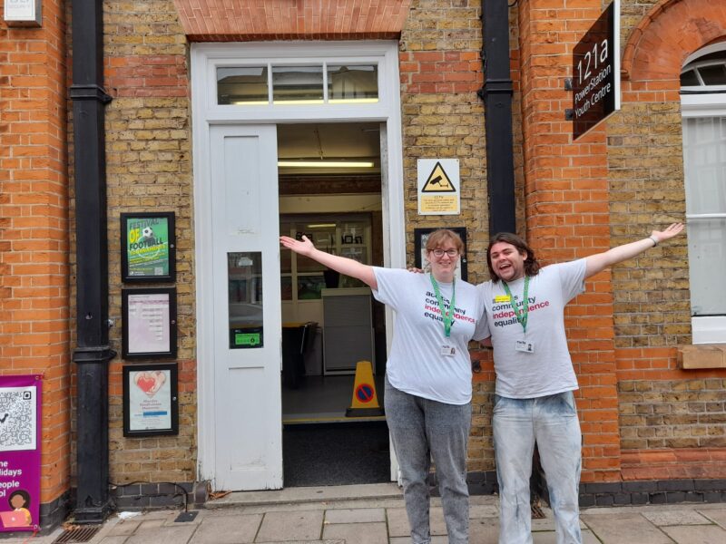 Image of Jenny and Ewan standing in front of the power station Youth centre ahead of a health and well being event. They are both wearing grey rules T shirts hidden disability sunflower lanyards and holding their arms out wide.