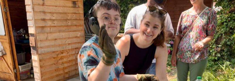 Image of a young man with Downs Syndrome waving at the camera with gardening gloves. There is a woman standing behind him supporting him to plant seeds.