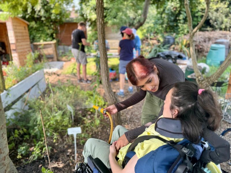 Image of a young woman's seated in a wheelchair watering the garden and an allotment with the support of her personal care assistant.