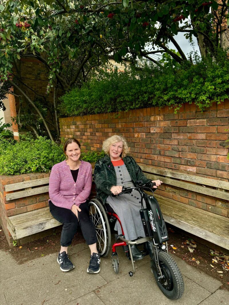 Our CEO Cathy and Chair of Trustees Jane in front of the Ruils office. Cathy is sitting on a wooden bench and Jane is in her wheelchair with a trike attachment.