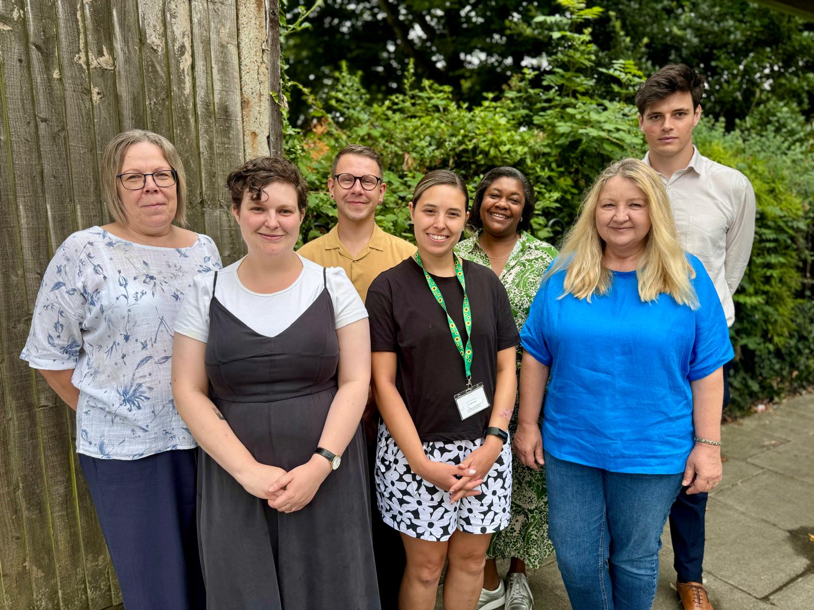 Group photo of the Proactive Anticipatory Care team and the Social Prescribing Manager. There are 7 of them and they are standing on the pavement with a wooden gate behind them.