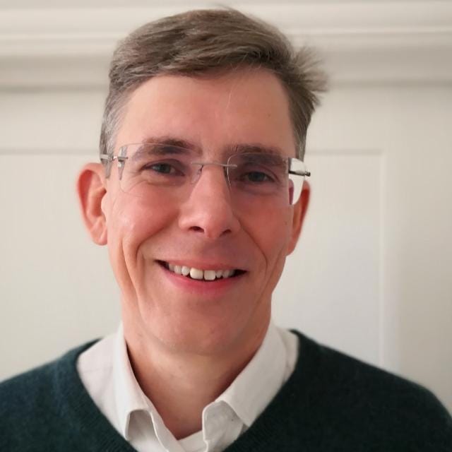 Image of a white man with light brown/grey short hair. He is wearing clear glasses and a navy jumper over a white collared shirt.