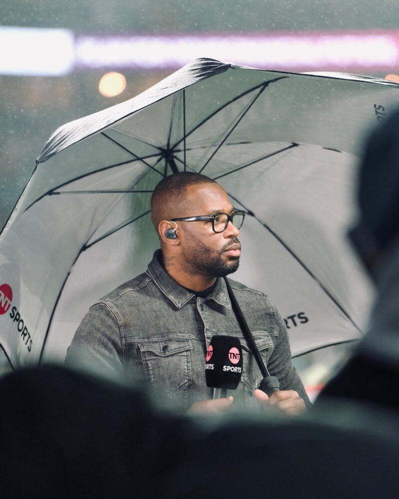 Image of Ugo Monye English sports pundit and former rugby union player standing under an umbrella with a microphone in his hand