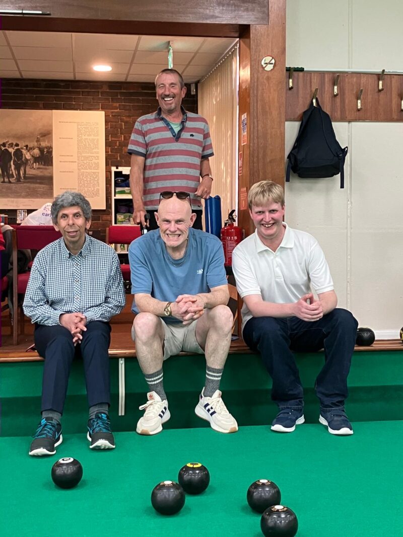 image of four people - 3 seated and one standing on an indoor bowls green