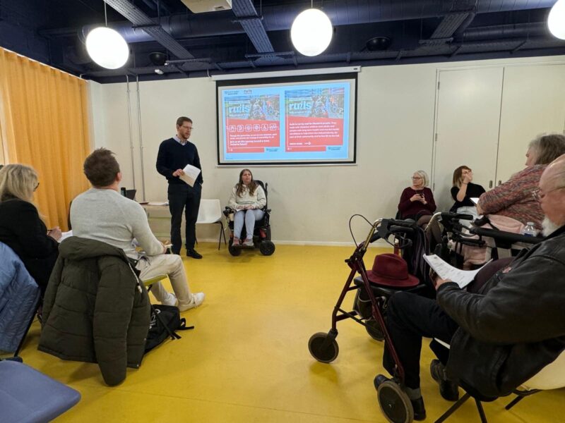 Image of a meeting room full of people facing a projector screen on the wall. The floor of the room is bright yellow and there are two people at the front giving a presentation.