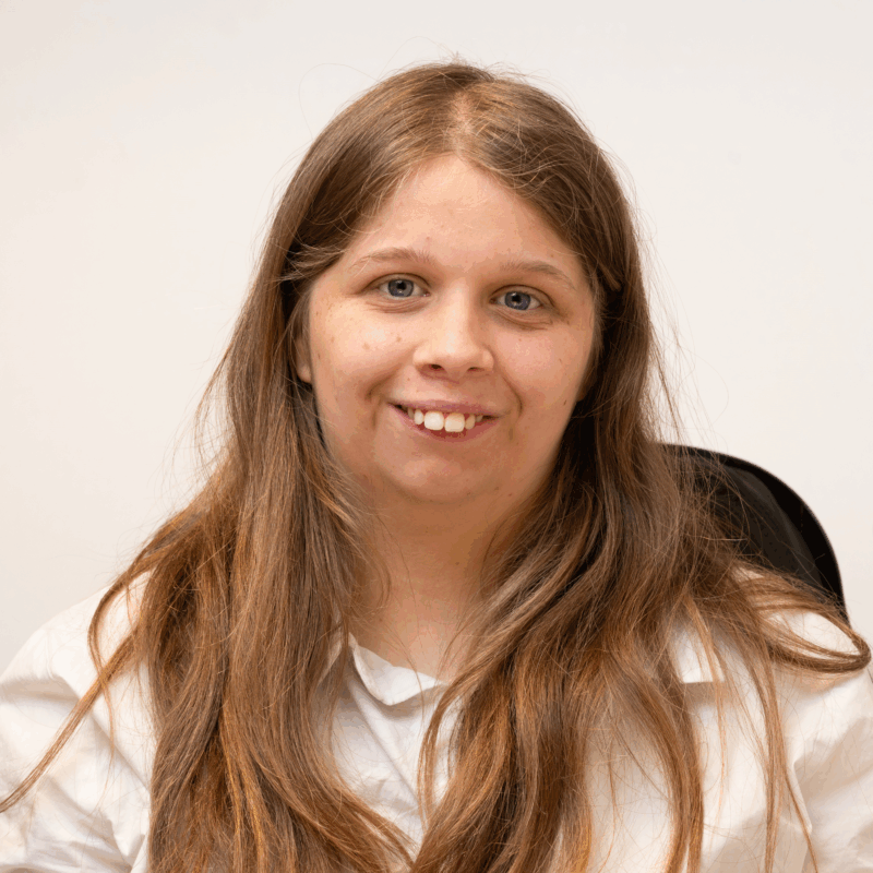 Image of a young woman with pale skin, dark blonde long hair and blue eyes. She is wearing a white shirt.