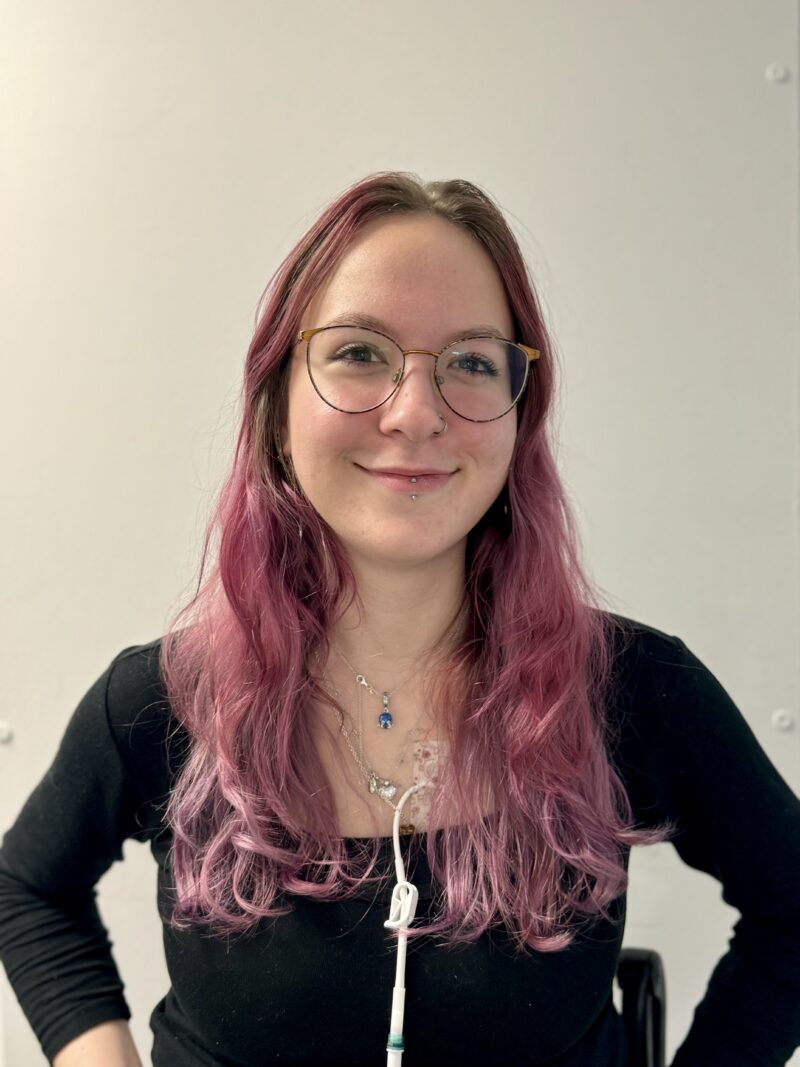 Woman with pale skin and brown and pink hair wearing glasses. She is wearing a black long sleeve shirt.