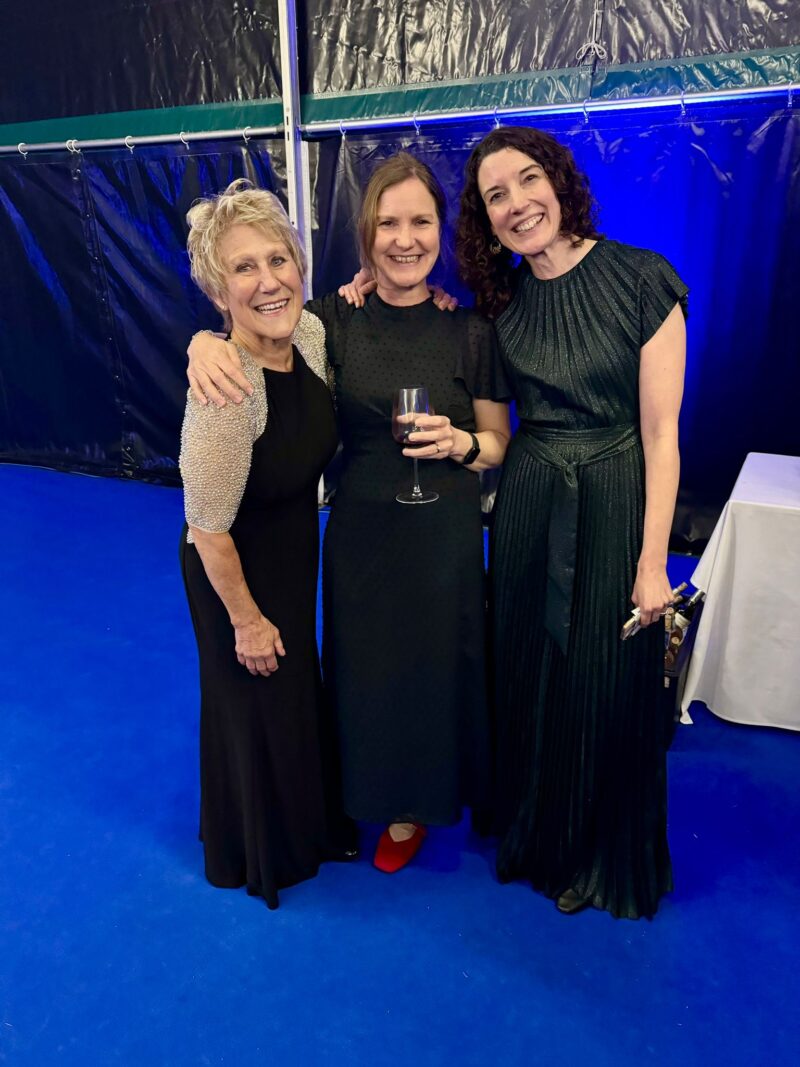 Image of three women standing in a event tent with blue carpet wearing formal black gowns