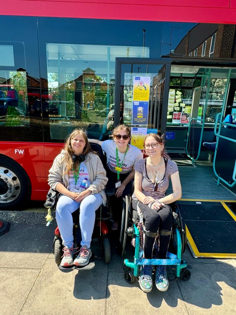 Image of three women in front of a red bus. Two are seated in their wheelchairs and the other is standing behind them.