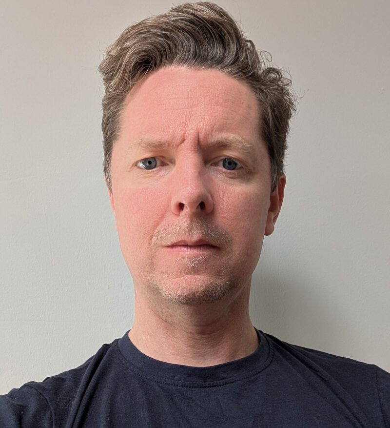 Image of a man with light brown and grey short hair with reddish pale skin, wearing a black t-shirt