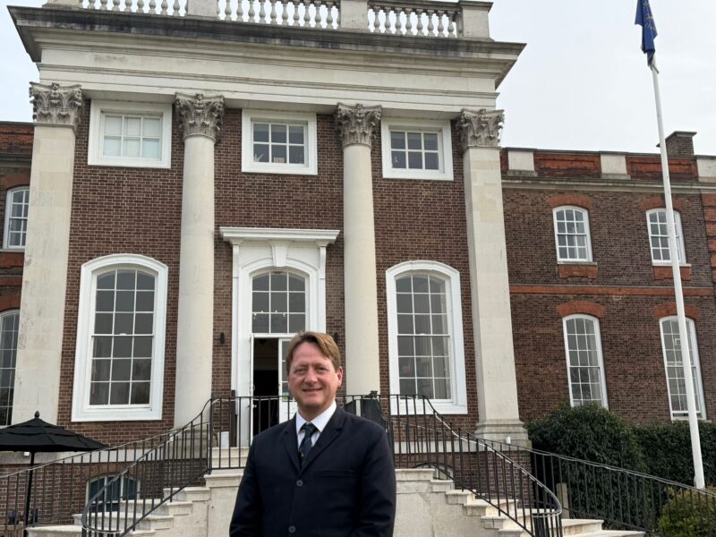 image of a man wearing a black suit standing in front of a gold club house