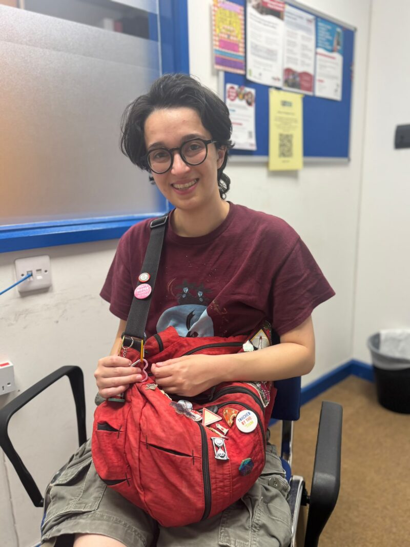 Image of a person sitting in a meeting room with a red bag with pin badges on it in their lap