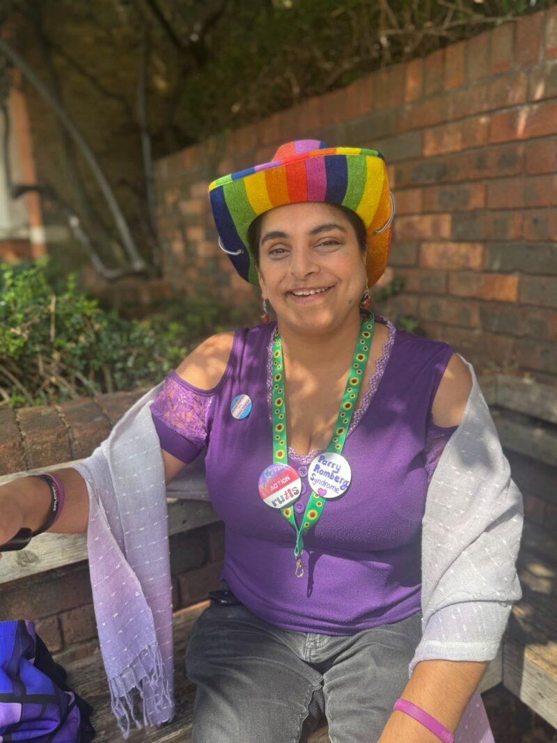 Image of a woman in a rainbow cowboy hat and purple top sitting with a walking stick on a bench