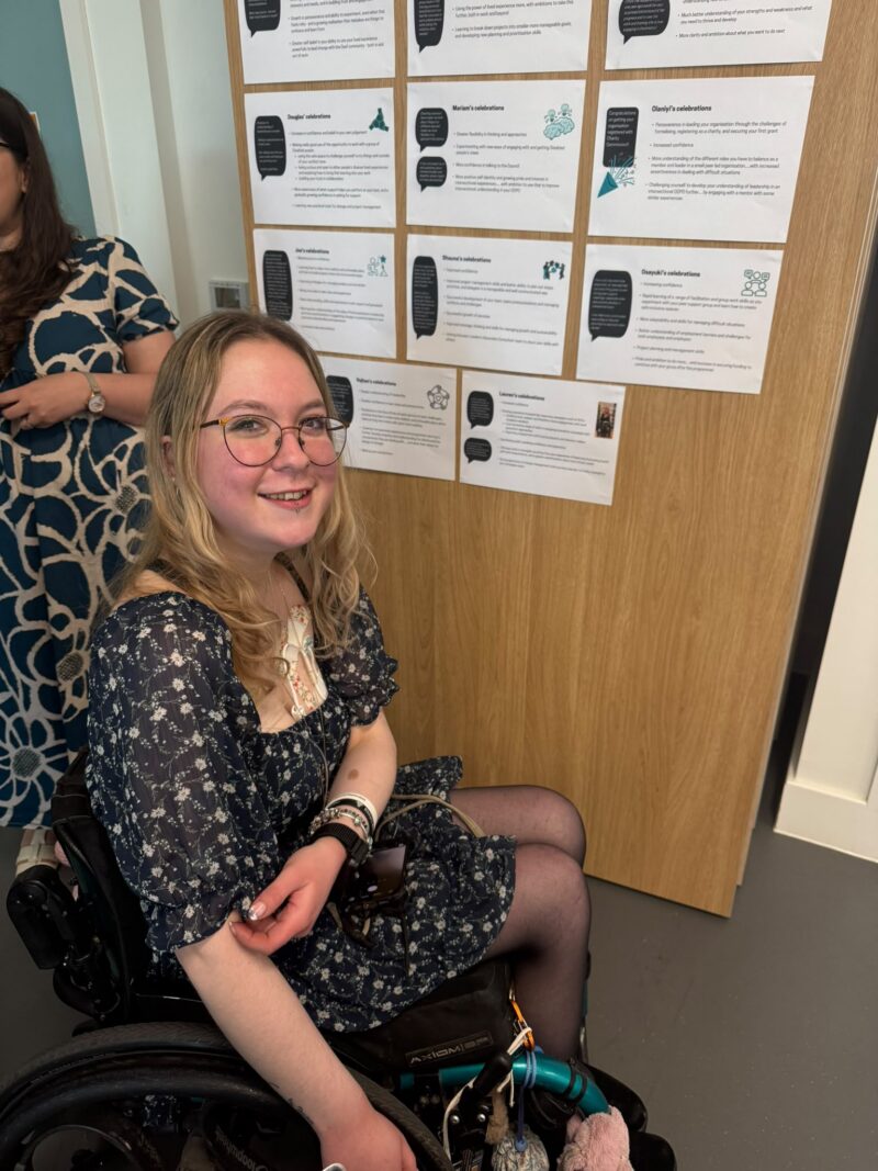 Image of a woman in a wheelchair sitting in front of a row of papers taped to a wall