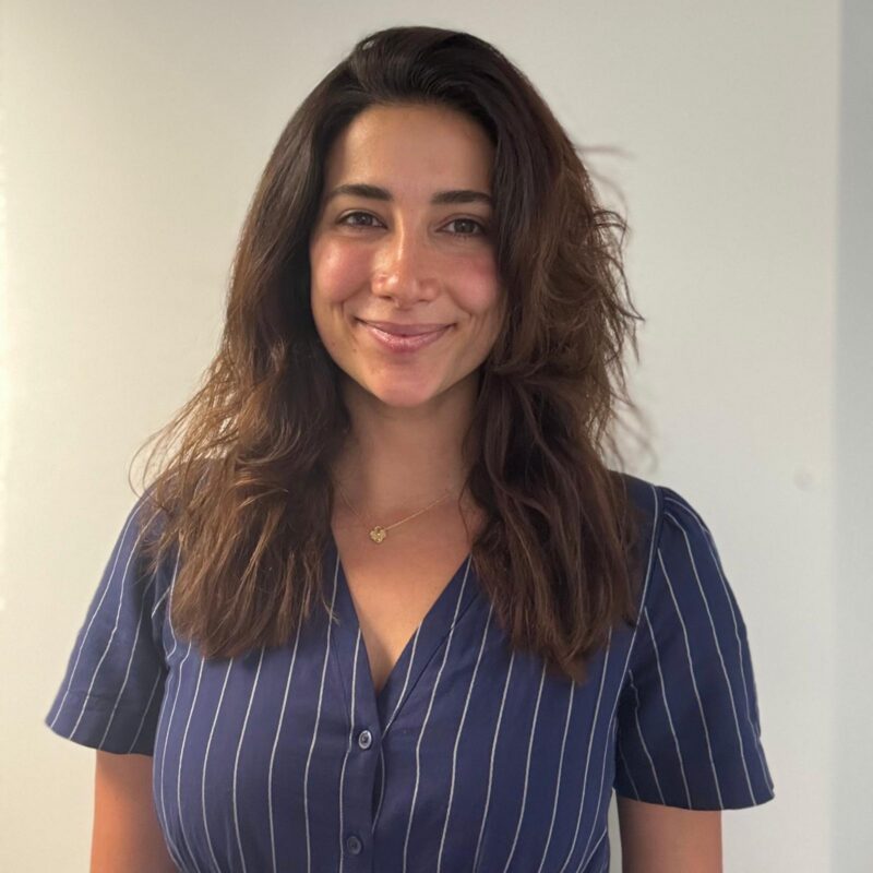 Image of a woman with light brown skin, medium brown chest length wavy hair, wearing a blue top with white stripes. She is smiling at the camera.